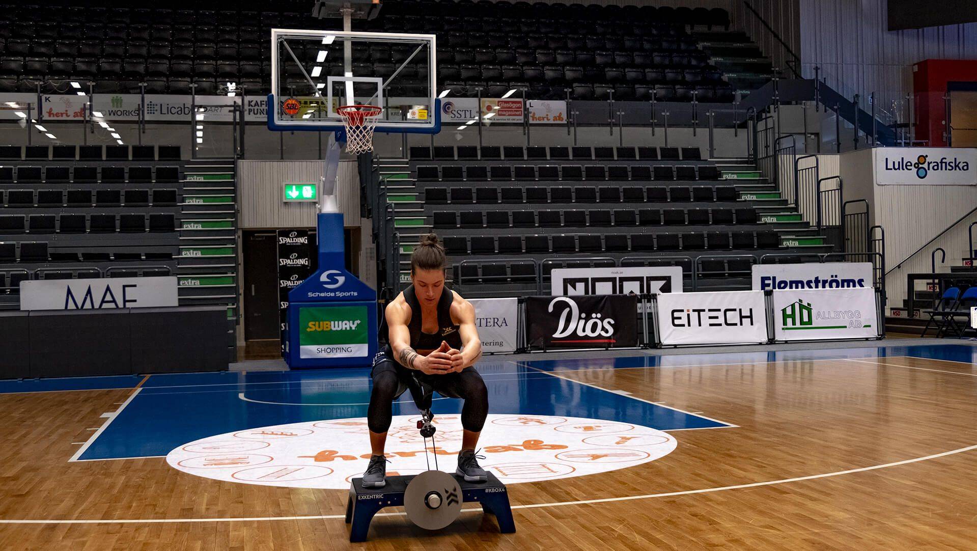 Maggie Lucas squatting on a kBox in a basketball arena