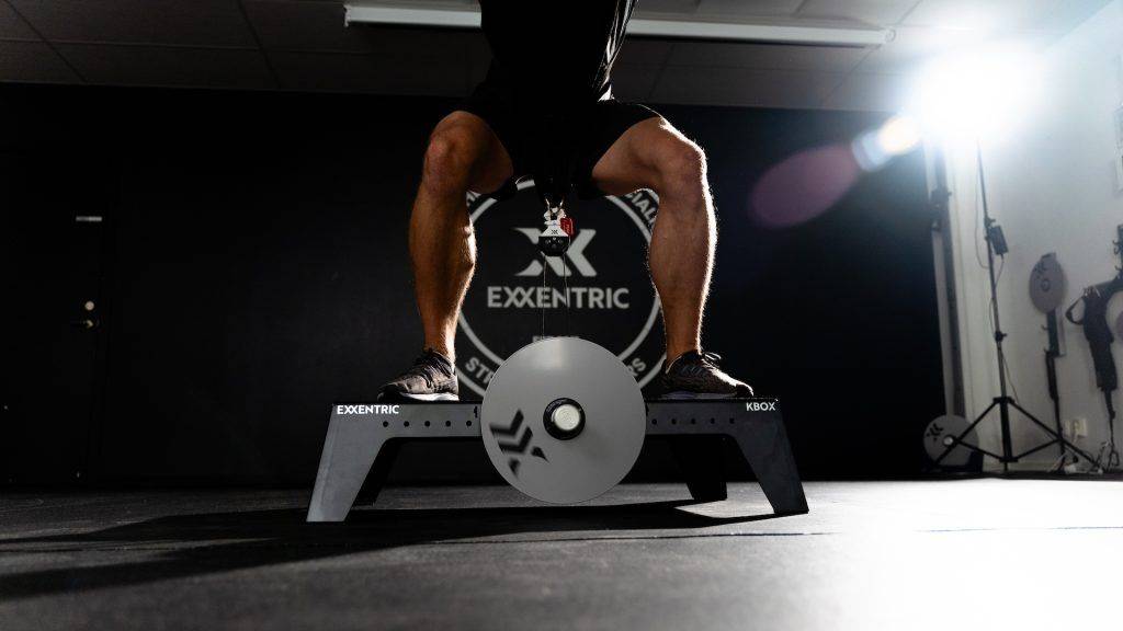 man squatting on kBox Pro with harness and L flywheel in a photo studio